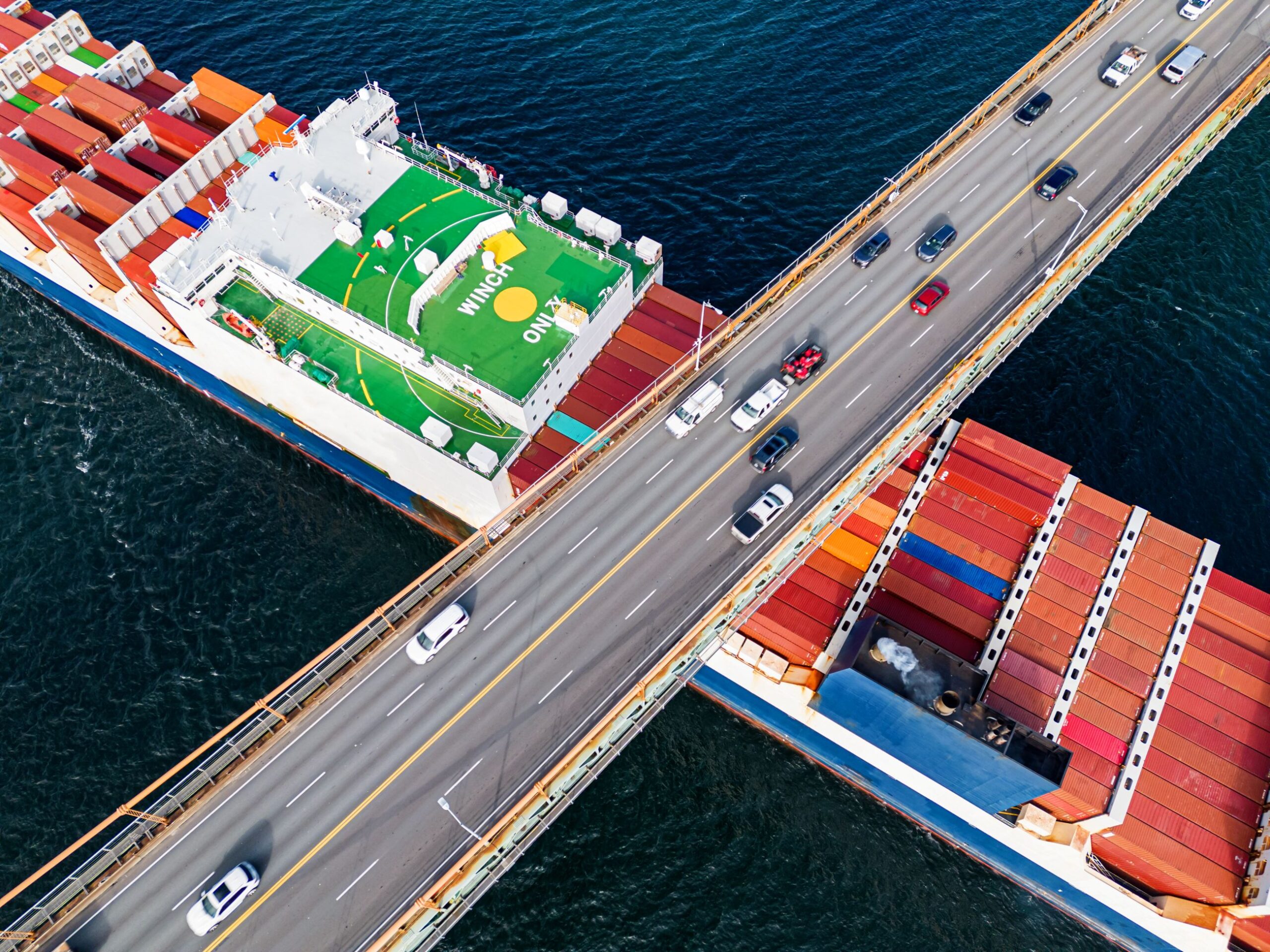 An aerial of a large ship carrying colourful shipping containers travelling under the MacKay Bridge. Cars travel in both directions on the bridge.