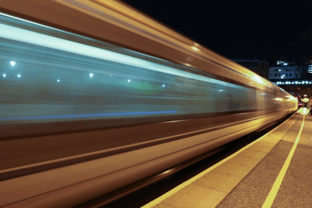 A train passes by a platform at high speed at night. The long exposure of the image stretches and distorts the train.